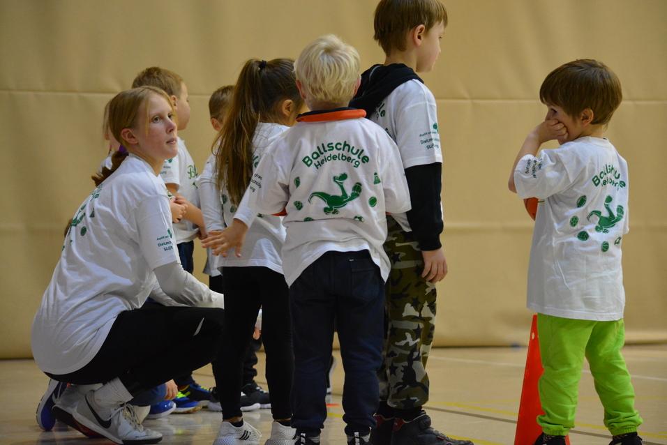 Eine Gruppe von Kindern in weißen T-Shirts steht mit einer Betreuerin in einer Sporthalle.