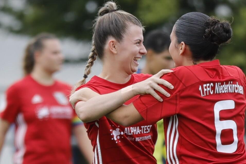 Zwei Fußballspielerinnen in roten Trikots umarmen sich lächelnd auf dem Spielfeld. Hintergrund unscharf.